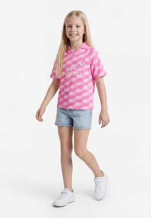 Uśmiechnięta dziewczyna idzie, ubrana w różową sportową koszulkę w kratę, jasne szorty jeansowe i białe trampki na jednolitym białym tle.