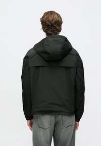 Άνδρας με σγουρά μαλλιά, φοράει μαύρο μπουφάν με κουκούλα και γκρι τζιν, γυρισμένος προς τα πίσω μπροστά σε έναν απλό φωτεινό φόντο.