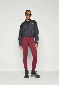 Črn skrajšan bomberski jopič z belimi črtami, rjavi legice in črne športne čevlje. Model nosi oversized sončna očala.