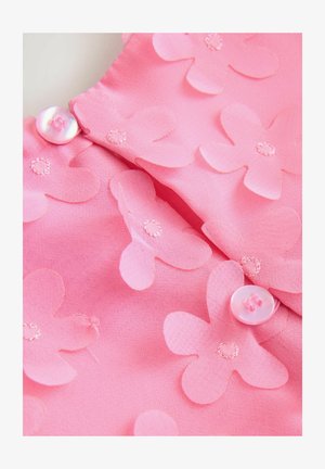 Rózsaszín szövet átfedő virágapplikációkkal, hímezett részletekkel és gombos záródással. Az anyag sima tapintású, finom mintázattal.