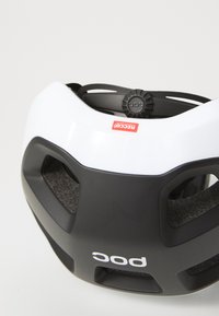 Tennisijal helmet, milleb detalliga kered, mustalt alumine sektsioon, ventilatsiooniaugud ning taga reguleeritav nupp sobivuse jaoks. Punased ja valged sildid nähtavad.