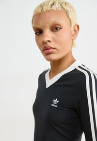 Camisola preta de manga longa com decote em V, com riscas brancas nos ombros e um logótipo da Adidas em branco bordado no peito. Material em mistura de algodão.