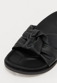 Black slide sandal with a knotted faux leather strap. Flat sole with a smooth texture and rounded toe. Minimalistic design.