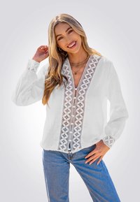 Witte blouse met verfijnde kantdetails aan de voorkant en manchetten, met een v-hals en lange mouwen. Gecombineerd met blauwe jeans.