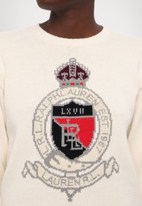 Gros plan sur un pull en tricot crème avec le blason Ralph Lauren comprenant une couronne, un écusson en noir et rouge, et le texte "R.L. Ralph Lauren EST 1967."