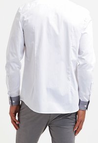 Camicia bianca a maniche lunghe con yoke posteriore, dettagli grigi ai polsini e una trama liscia, abbinata a pantaloni grigi chiari.
