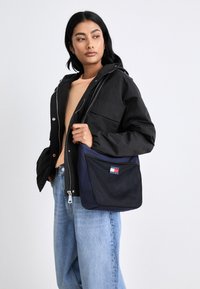 Navy fabric shoulder bag with a black mesh front pocket, featuring a red, white, and blue logo. Smooth texture and rectangular shape.