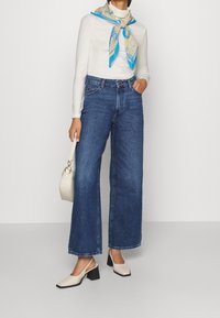 Hohe, weit geschnittene blaue Jeans kombiniert mit einem taillierten weißen Langarmshirt und einem hellblauen, gemusterten Schal. Weiße Blockabsatzschuhe.