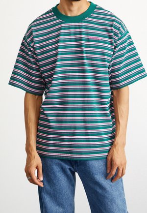 Mężczyzna ubrany w luźną koszulę w paski w kolorach turkusowym, różowym i białym z krótkim rękawem oraz niebieskie dżinsy, stojący na białym tle.