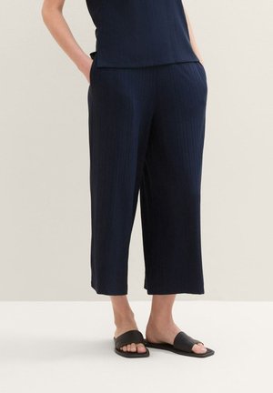 Osoba nosząca granatowe skrócone spodnie o szerokich nogawkach oraz czarne klapki, stojąca z rękami w kieszeniach na neutralnym tle.