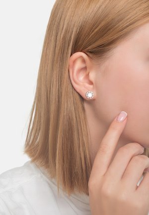Runda stiftörhängen i silverfärgad metall med en geometrisk utskärningsdesign. Modellen har rakt, axellångt blont hår.