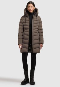 Ruskea puhvitakki, jossa on huppu, pystysuora tikattu kuvio, vetoketju edessä ja sivutaskut. Päällä mustat legginsit ja paksut saappaat.