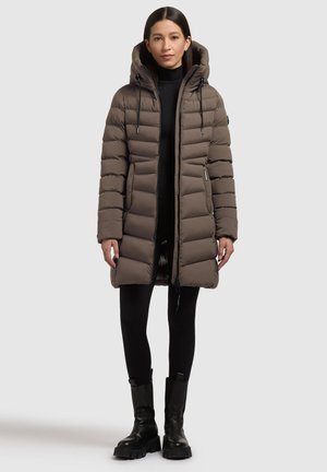 Abrigo puffer marrón con capucha, con un patrón de acolchado vertical, cremallera frontal y bolsillos laterales. Llevado sobre leggings negros y botas gruesas.