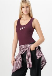 Burgundy tank top with "GAP" logo; ribbed texture. Paired with a pink and black checkered flannel tied around the waist. Black pants.