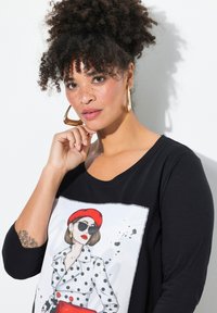 Schwarzes Langarmshirt mit einem Grafikdesign einer Frau in gepunktetem Kleid und rotem Hut. Das Material sieht glatt aus und hat eine lockere Passform.