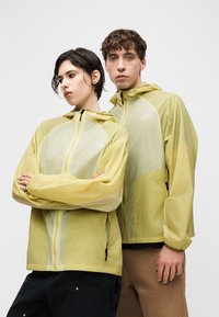 Deux jeunes adultes portant des vestes à capuche jaune pâle, debout près l'un de l'autre avec des expressions neutres sur un fond blanc uni.