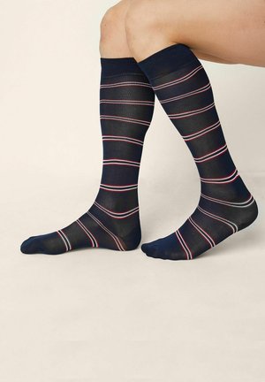 Legs wearing navy blue knee-high socks with thin white and red horizontal stripes, positioned against a plain beige background.