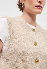 Bežna teksturirana brezrokavnik izdelana iz puhastega materiala, z okroglim ovratnikom in tremi zlatimi gumbi spredaj. Nosi se čez belo srajco.