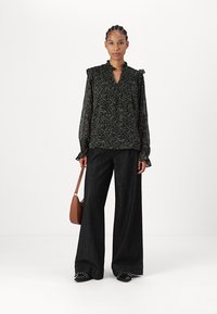 Svart bluse med blomstermønster, volangkrage; kombinert med mørke, vide jeans; modellen har en brun veske; svarte sko med detaljer.