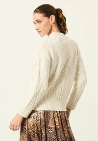 Grobmaschiger, cremefarbener Strickpullover mit Rautenmuster, geripptem Saum und langen Ärmeln, kombiniert mit einem Rock aus seidigem Leopardenmuster.