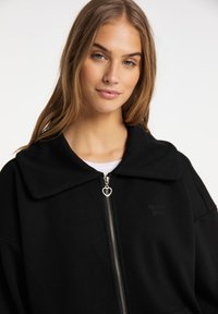 Giacca nera con zip e colletto ampio, dotata di una zip con tirante a forma di cuore e un delicato dettaglio del logo. Tessuto morbido con una vestibilità comoda.