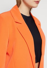 Oranžový blejzr se strukturovaným designem, s klopami a volným střihem, kombinovaný s černým kratším topem pod ním.