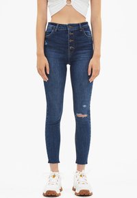 Högmidjade, mörkblå jeans i denim med knappsluiting, smal passform och ett slitage på höger knä. Stretchigt tyg med lätt blekning.