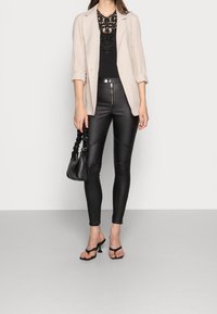 Frau trägt eine beige karierte Blazerjacke, ein schwarzes Spitzenoberteil, schwarze Lederhosen, eine schwarze geraffte Handtasche und schwarze Absatzsandalen vor einem einfarbigen Hintergrund.