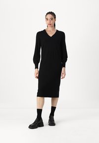 Juoda megzta suknelė su V formos iškirpte ir ilgomis burbulinėmis rankovėmis, iki kelių, pasižyminti prigludusia siluetu ir lygiu paviršiumi.