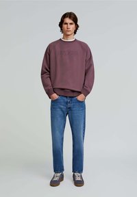 Felpa a girocollo in un viola tenue con logo "SCALPERS" in rilievo, abbinata a jeans dritti blu medio e sneakers grigie.