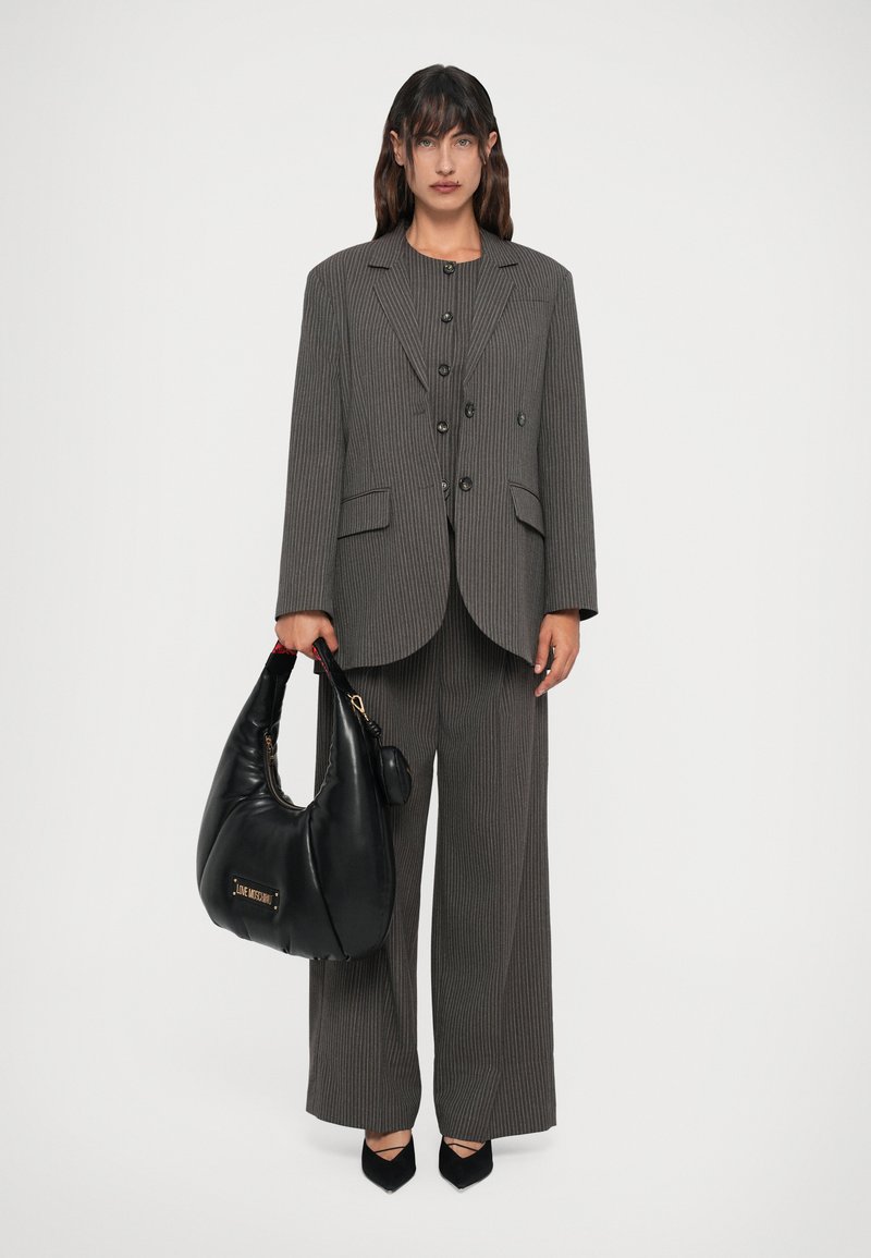 Grau gestreifter Blazer und weite Hose mit schwarzen Akzenten. Das Model hält eine große schwarze Tasche mit einem goldenen Logo. Spitz zulaufende schwarze Schuhe an den Füßen.
