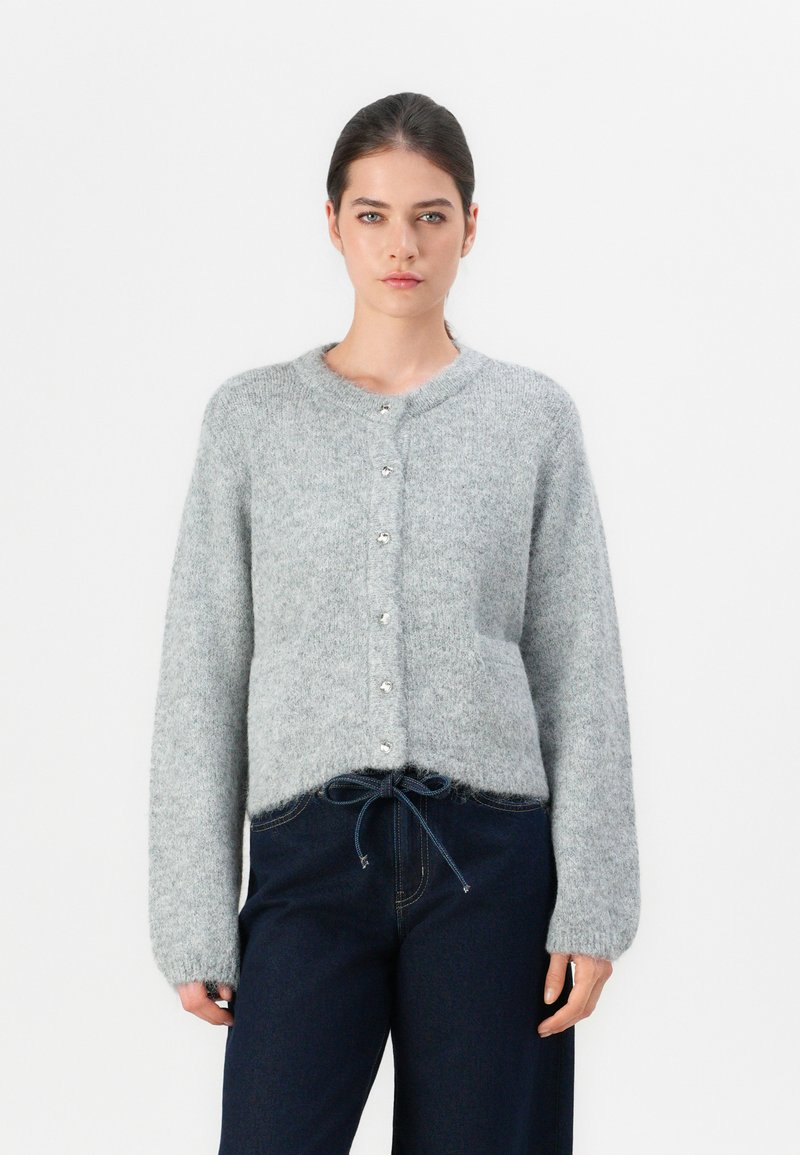 Grijze gebreide cardigan met een ronde halslijn, knoopsluiting aan de voorkant en een zachte textuur. Beschikt over een zak en een trekkoorddetail aan de zoom.