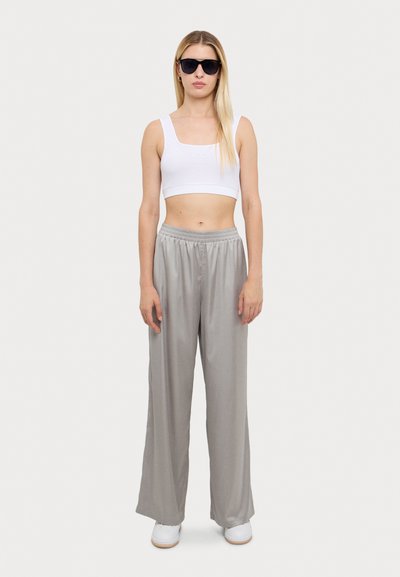 Lys grå joggebukser med bred benvidde og glatt tekstur, sammen med en tettsittende hvit crop top og svarte solbriller. Hvite sneakers fullfører antrekket.