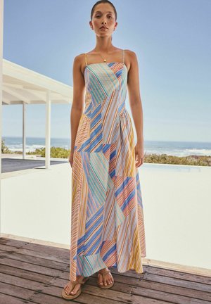 Naine seisab puidust terrassil basseini ääres, kannab varrukateta mitmevärvilise triibulise pisipalliga maksikleit koos õhukeste rihmade ja sandaalidega.