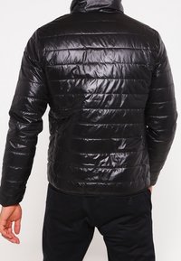 Black, shiny puffer jacket viewed from the back, featuring a quilted pattern, high collar, and fitted silhouette. Warm, lightweight fabric.
