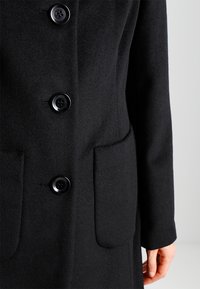 Zwarte wollen jas met drie grote zwarte knopen, twee voorkruiken en een hand van een persoon zichtbaar aan de zijkant.