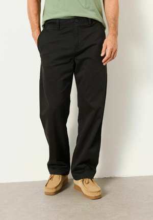 Mann trägt schwarze Hose mit geradem Bein, braune Wildlederschnürschuhe und ein grünes Hemd, steht mit den Händen in den Taschen auf grauem Boden.