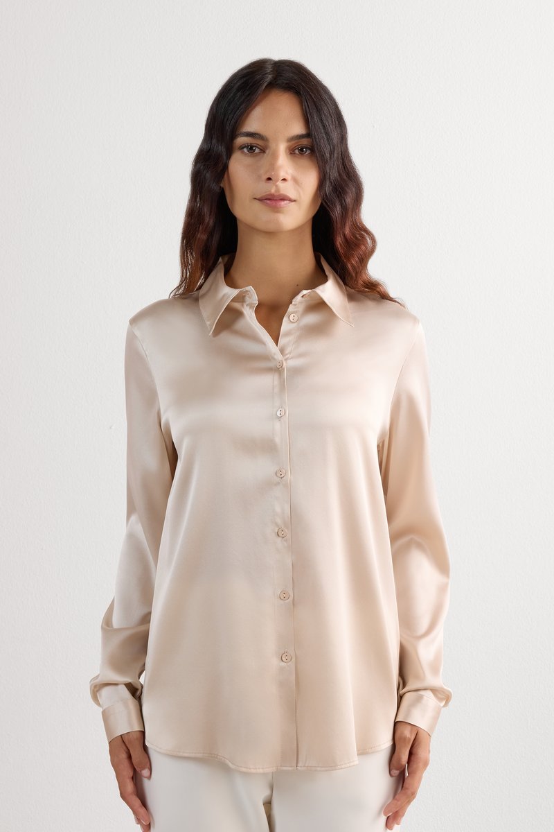 Kvinde iført beige satin langærmet skjorte med knapper og spids krave, stående mod en lys, ens baggrund.