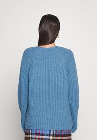 Sininen neulepaita, jossa on ribbikudos, raglan-hihat ja pyöreä kaula-aukko. Paita on päällä ruutuhameen, ja siinä näkyy takapuolen muotoilu sekä ompeluyksityiskohdat.