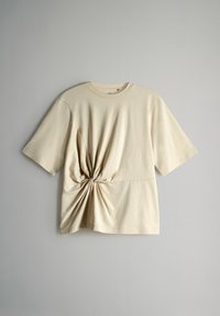 Bež lühikeste varrukatega t-särk, mis on valmistatud pehmest kangast. Omab keeratud sõlme detaili vööl ja lõdvat istuvust. Sile tekstuur ja ümmargune kaelus.