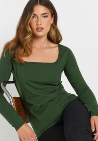 Langärmliges grünes Oberteil mit quadratischem Ausschnitt, weicher Textur und taillierter Silhouette, kombiniert mit schwarzen Hosen.