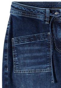Gros plan sur un jean en denim bleu foncé montrant la poche avant, les passants de ceinture, le bouton et les détails de couture visibles.