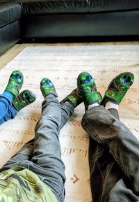 Groene klompen met zwarte accenten hebben een levendig ontwerp van een kikker. gedragen met grijze jeans op een gemêleerd tapijt, dat textuur en kleurcontrast laat zien.