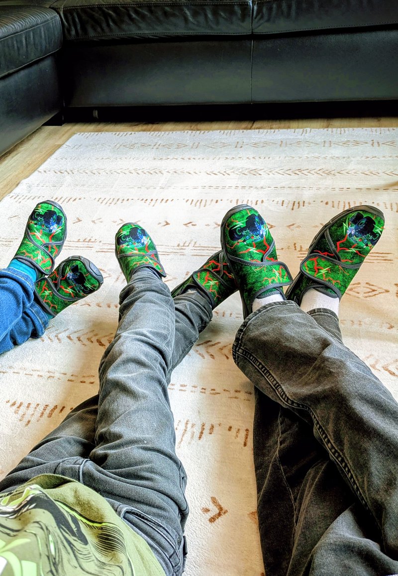 Groene klompen met zwarte accenten hebben een levendig ontwerp van een kikker. gedragen met grijze jeans op een gemêleerd tapijt, dat textuur en kleurcontrast laat zien.