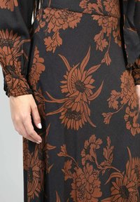 Robe en tissu noir avec un motif floral marron, présentant des détails de manches froncées et une texture lisse. Vue en gros plan du design et de la main.