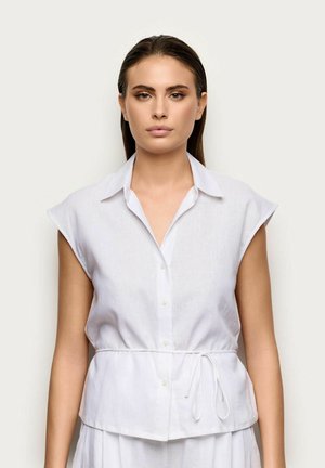 Donna con capelli castani lisci che indossa un top bianco senza maniche con bottoni, colletto e cintura legata in vita, in piedi davanti a uno sfondo uniforme.