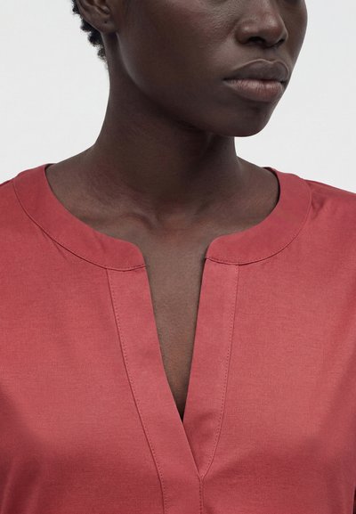 Κοντινό πλάνο ενός ατόμου που φοράει ένα ροδοκόκκινο μπλουζάκι V-λαιμόκοψη με στρογγυλή λαιμόκοψη, εμφανίζοντας τον λαιμό και το κάτω μέρος του προσώπου σε απλό φόντο.