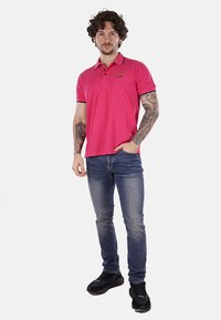 Polo shirt rosa con dettagli scuri sul colletto, abbinato a jeans blu. Il modello è in una pose neutra, mostrando la vestibilità e il colore della camicia.