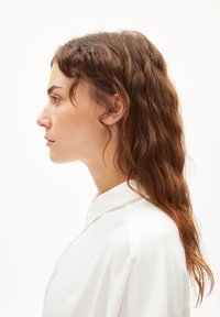 White, collared blouse with a smooth texture, featuring a relaxed fit. Model has long, wavy brown hair, viewed from the side.