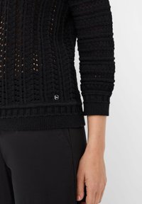 Schwarzer Strickpullover mit strukturierten Mustern, ausgestattet mit gerippten Bündchen und Saum. Enthält ein kleines metallisches Logo-Element am unteren Ende.